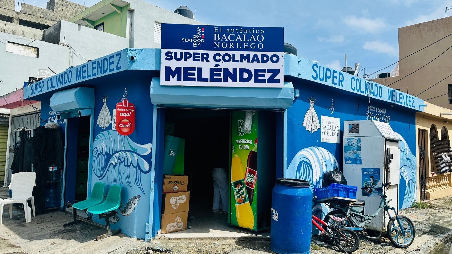 En blåmalt liten butikk som heter Super Colmado Melénez. Ulik reklame rundt butikken, deriblant Seafood from Norway og El auténtico Bacalao Noruego